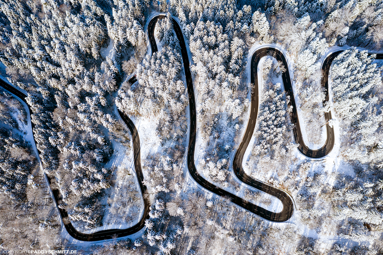 Kurvige Landstraße aus der Vogelperspektive die durch einen schneebedeckten Wald im Allgaeu führt.