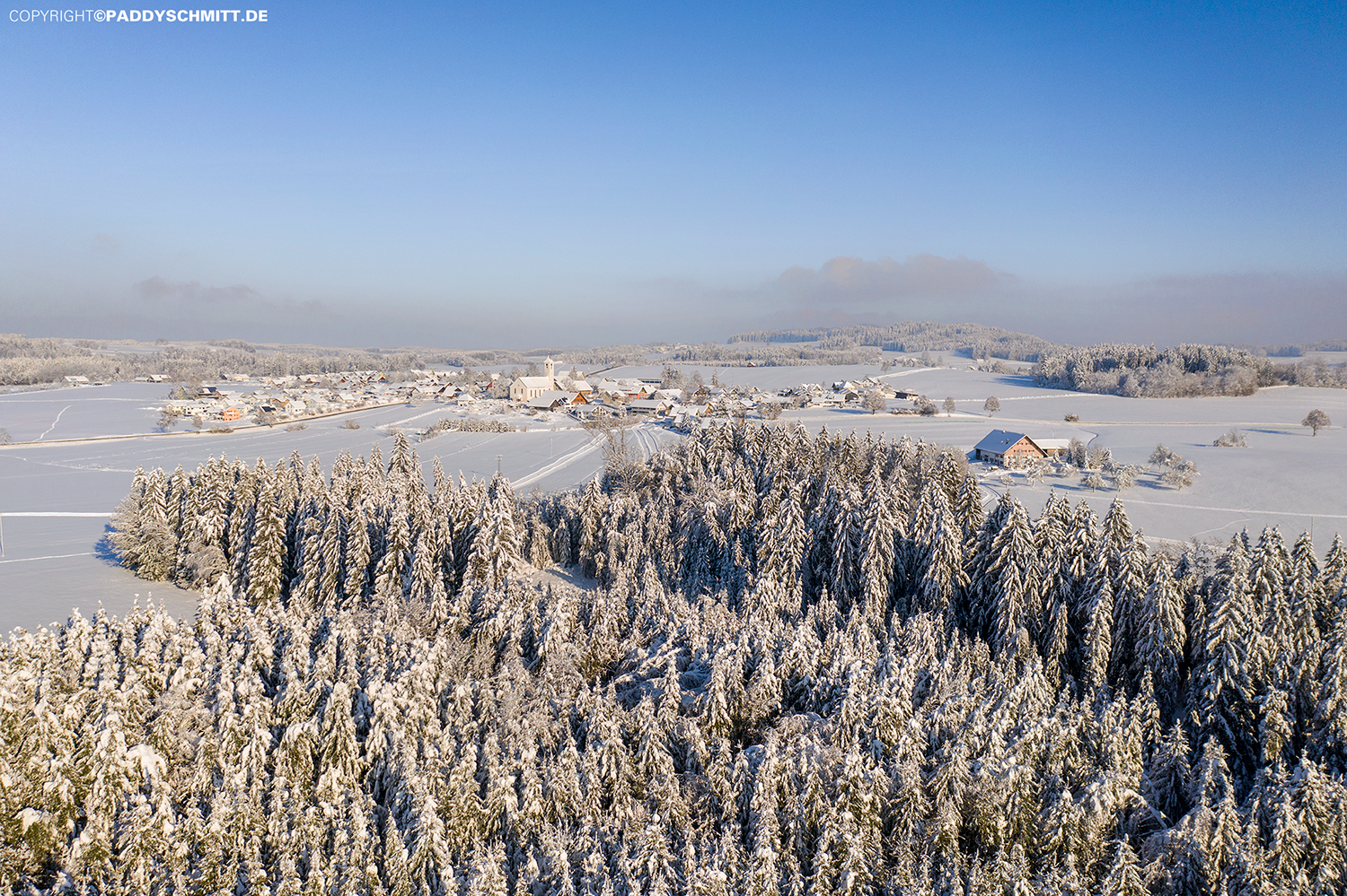 Allgaeuer Landschaft im Winter mit einem Wald und einem kleinen Dorf.